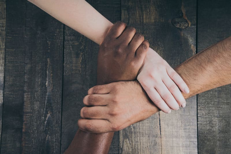 Three people of different skin tones holding one another by the wrist.